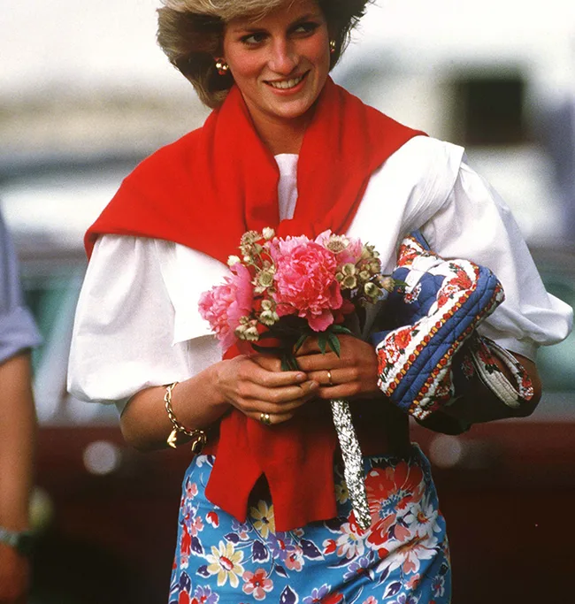 Woman in a floral skirt and red scarf, holding a bouquet and smiling outdoors.