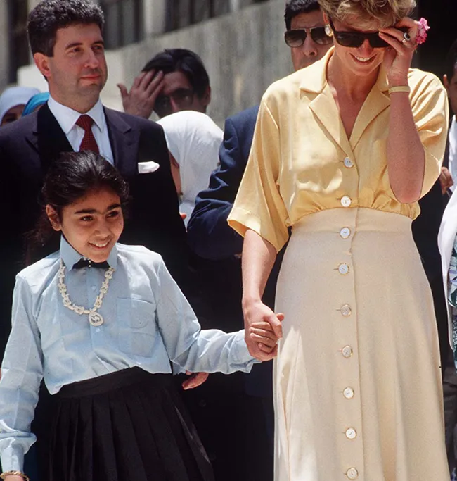 A woman in a yellow outfit holds the hand of a young girl in a blue shirt, walking outside with people in the background.