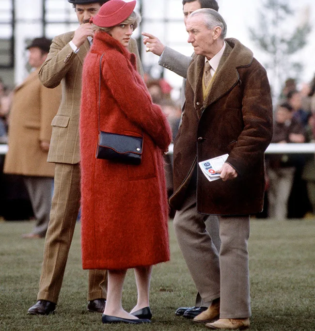 A woman in a red coat and hat stands on grass among people dressed for a formal event, with a man gesturing beside her.