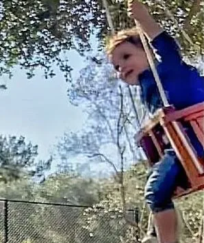 Toddler in blue shirt swings joyfully outdoors, surrounded by trees and a clear sky.