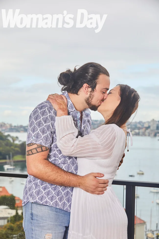 A couple kisses on a balcony with a scenic waterfront backdrop, featured in Woman's Day.