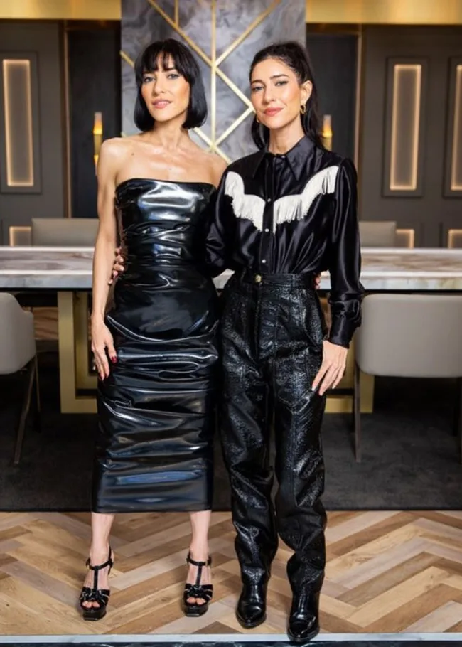 Two women in stylish black outfits pose together in a modern room with wood flooring and elegant decor.
