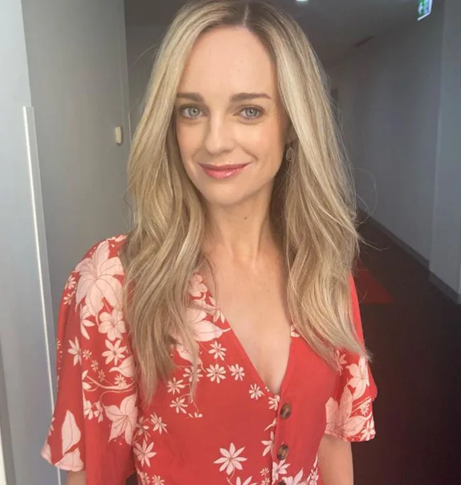 A woman with long blonde hair wearing a red floral dress smiles in a hallway.