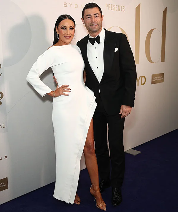A woman in a white dress and a man in a tuxedo pose together on a red carpet against a branded backdrop.