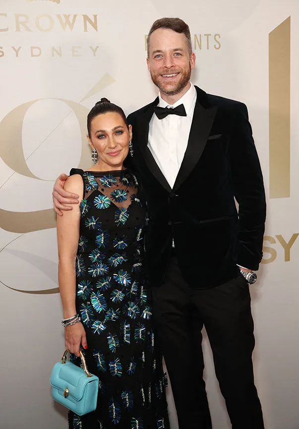 A couple poses on the red carpet at a formal event, with the woman in a floral dress and the man in a tuxedo.