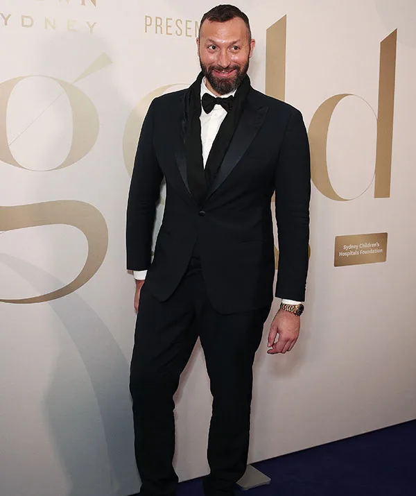 Man in a black suit and bow tie at a formal event with a gold-themed backdrop.