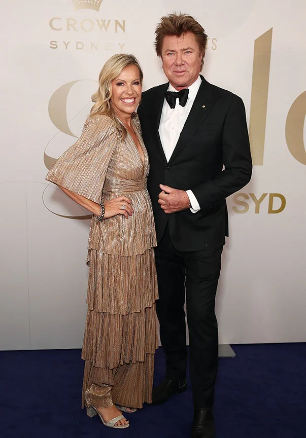 A man in a tuxedo and a woman in a gold gown smile on a blue carpet at a formal event in Sydney.