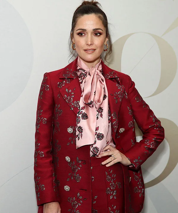 A person in a red floral suit with a pink bow blouse poses against a neutral background.