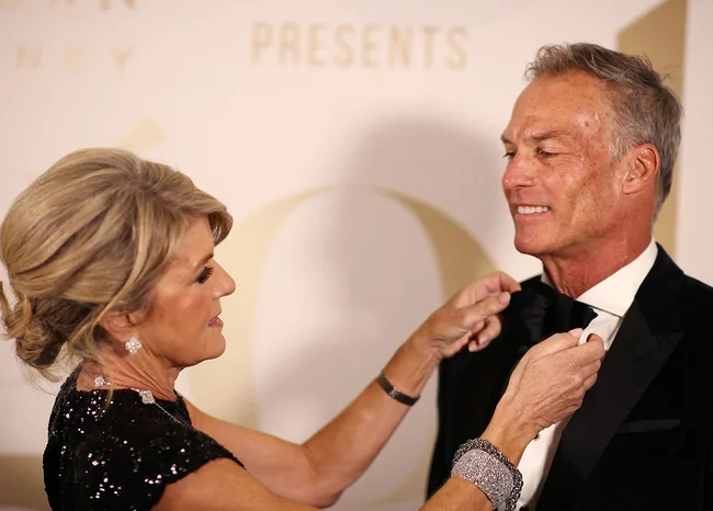 A woman adjusts a man's bow tie at a formal event. Both are smiling.