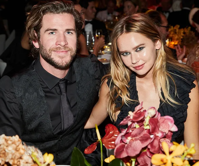 Man and woman in formal attire sitting at a table with flowers, smiling at a formal event.
