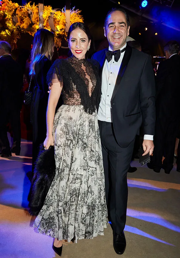 Elegant couple in formal attire at an upscale event, with the man in a tuxedo and the woman in a lace and floral gown.