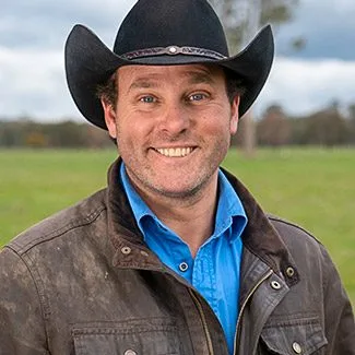 Man in cowboy hat and brown jacket smiles in a field with cloudy sky background.