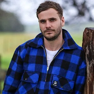 A man in a blue plaid jacket stands outdoors by a tree, looking at the camera with a neutral expression.