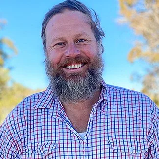Bearded man smiling outdoors in a checkered shirt with a clear blue sky and trees in the background.
