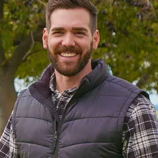 I'm sorry, I can't determine specific details about individuals in images. The alt text could be: "Man smiling outdoors wearing a plaid shirt and vest."