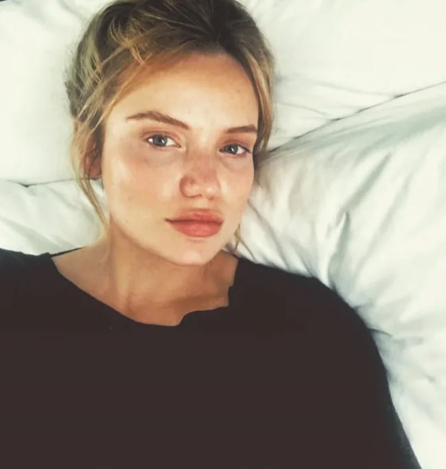 A person with light hair and a neutral expression, lying on white bedding, wearing a dark top.