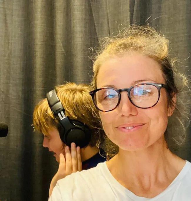Woman with glasses smiles at camera; child with headphones in background.