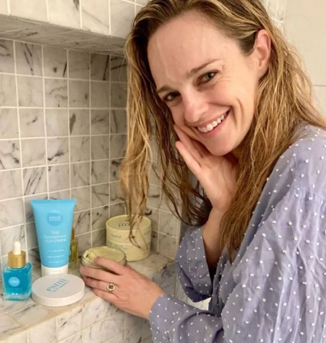 Woman in polka dot robe smiles near skincare products on a tiled bathroom shelf.