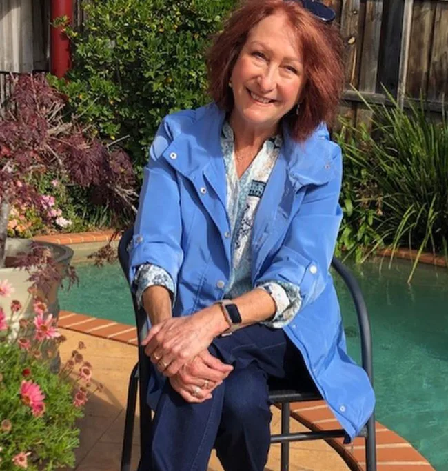 Woman in blue jacket sitting by a pool, smiling, with garden plants around.
