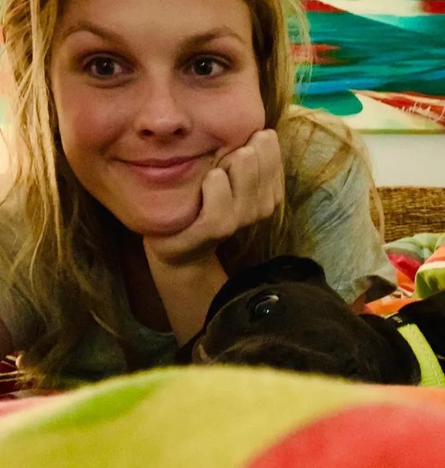 A woman smiling with her head resting on her hand, alongside a black dog on a colorful blanket.