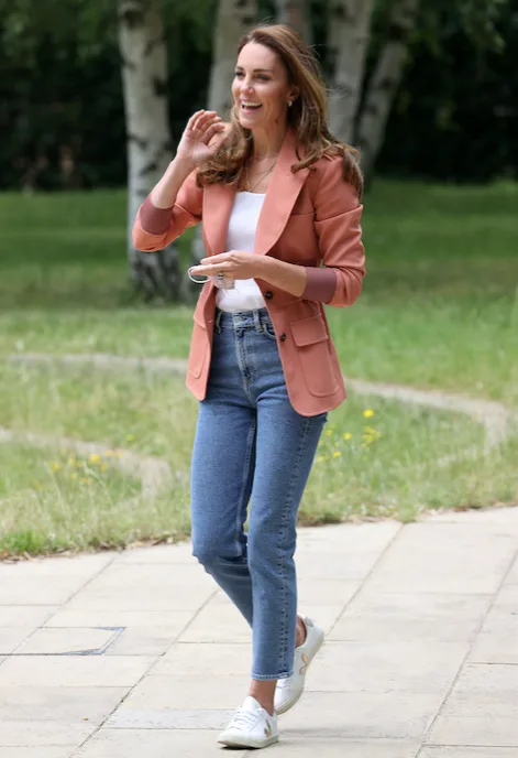A woman outdoors in a pink blazer, jeans, and white sneakers, smiling and holding sunglasses.