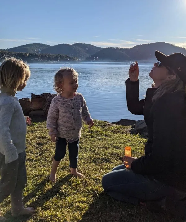 A person blows bubbles by a lake with two children nearby, under a clear blue sky with mountains in the background.