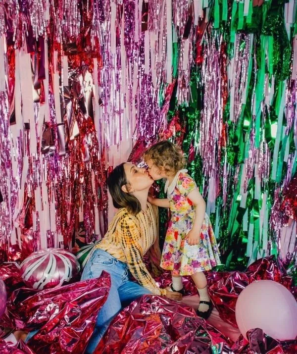 Woman and child share a kiss in a colorful backdrop with shiny pink and green streamers and balloons.