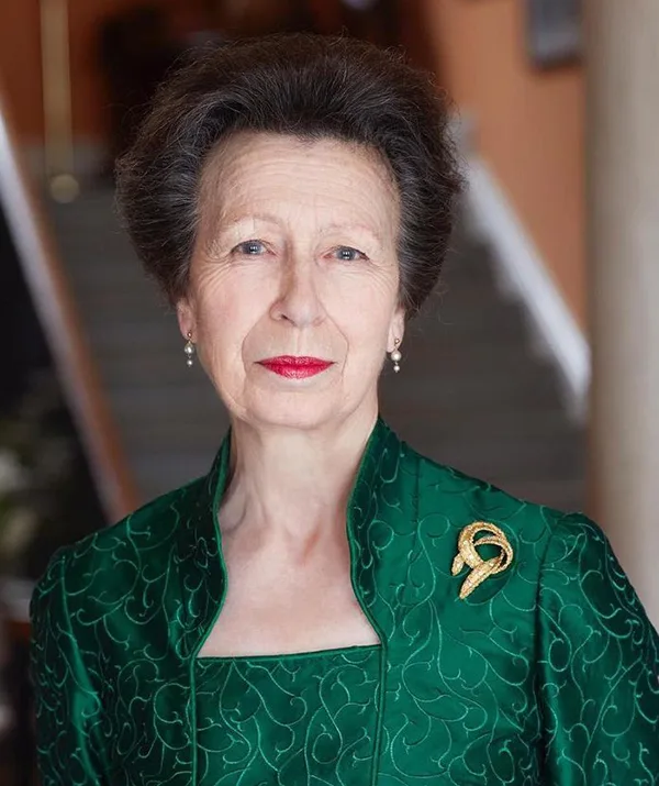 Woman in elegant green dress with ornate brooch stands indoors with a graceful expression, wearing pearl earrings.