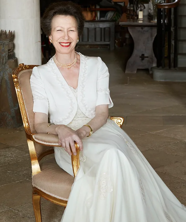 A woman in elegant white attire sits on a gold-trimmed chair, smiling warmly in a well-appointed room.