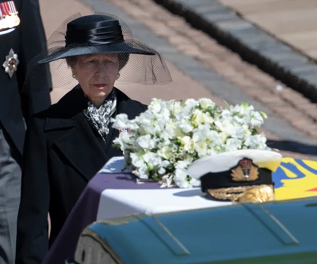A woman in black attire and hat stands near a flower-covered casket draped with a flag bearing a naval hat.