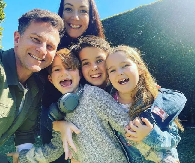 Family of five smiling outdoors, with two adults and three children embracing each other.