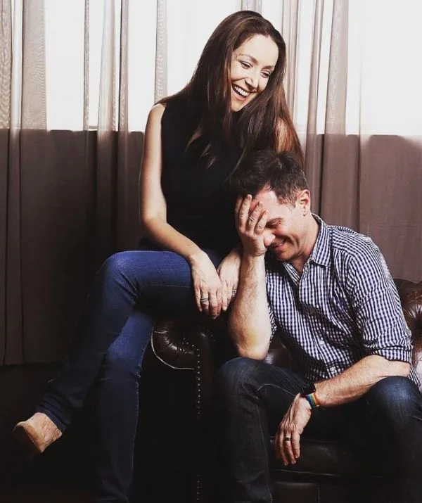 A woman and a man smile and sit together on a couch, with soft lighting and curtains in the background.