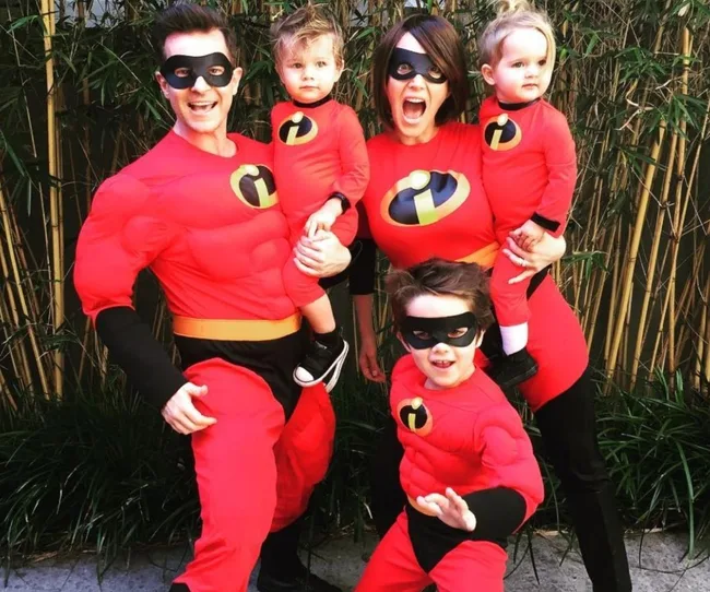 Family dressed in matching "Incredibles" superhero costumes with black masks, posing outdoors.