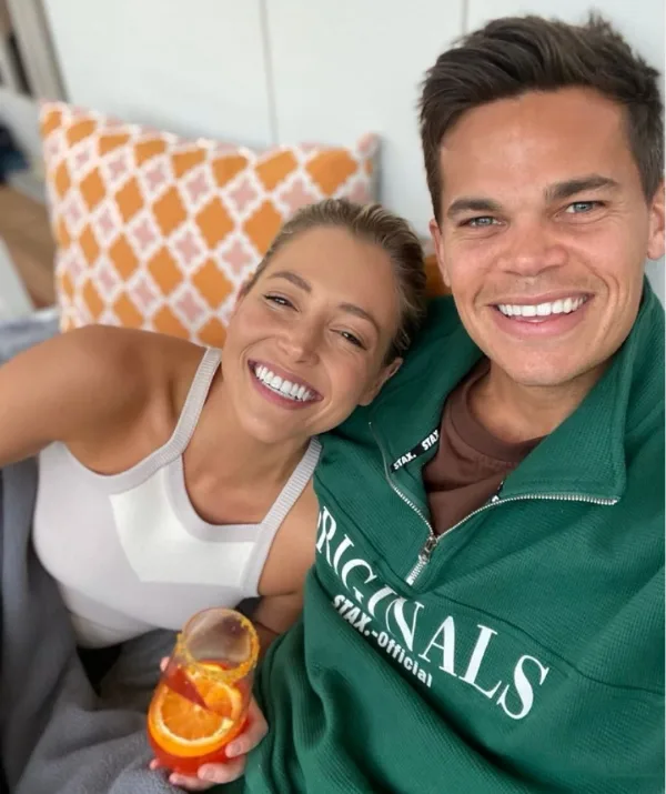 A smiling couple sitting close, she holds a drink with an orange slice; background shows an orange and white pillow.