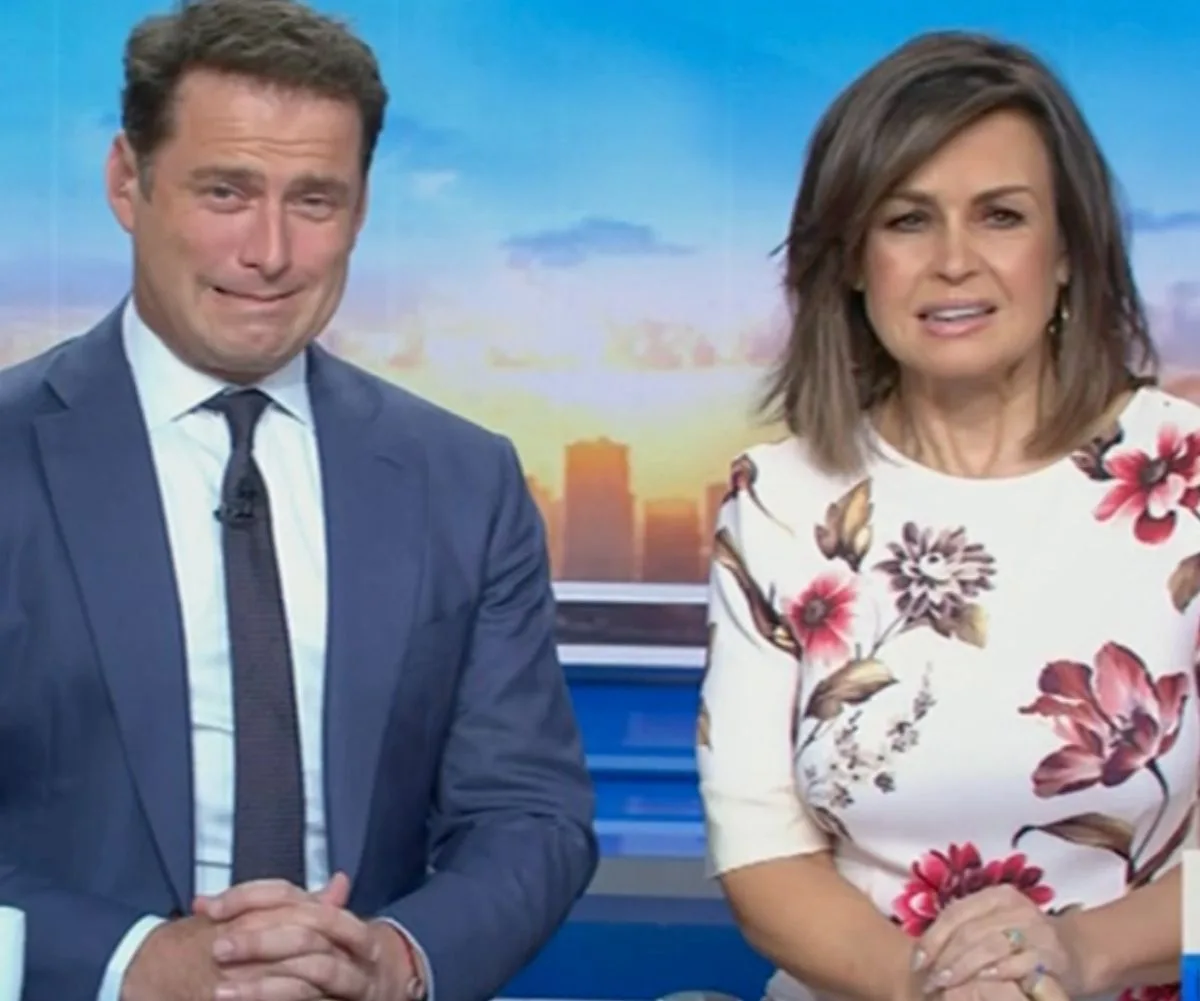 Two TV hosts sitting at a desk during a live broadcast, one in a suit and the other in a floral dress.