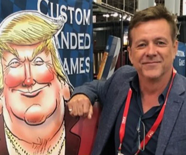 A man poses with a cartoon standee of a smiling, blond-haired figure at a custom branded games booth.