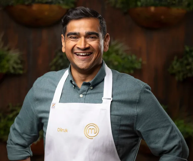 Dilruk in a MasterChef apron smiling, standing in front of a dark, plant-decorated background.