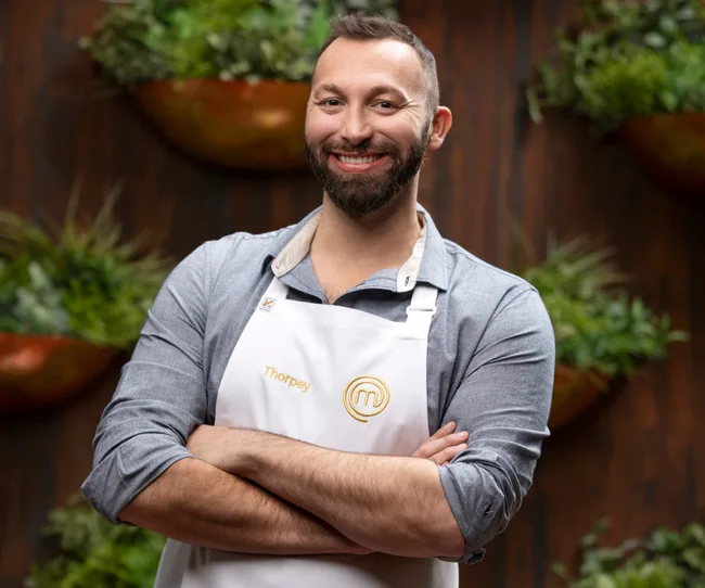 A person smiling, wearing a white apron with "Thorpey" on it, standing with arms crossed in front of a plant wall.
