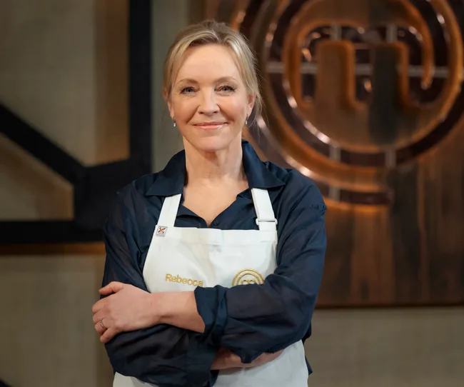 A person in a "MasterChef" setting, wearing a white apron with "Rebecca" embroidered, stands smiling with arms crossed.