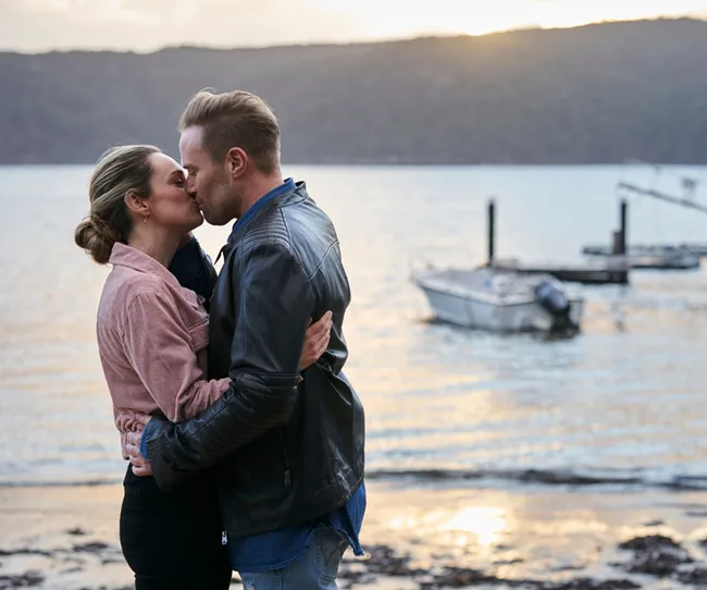 A couple kissing by the sea at sunset, with boats on the water in the background.