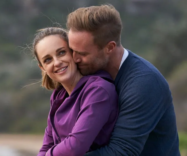 A couple embraces outdoors, smiling warmly at each other, with a blurred natural background.