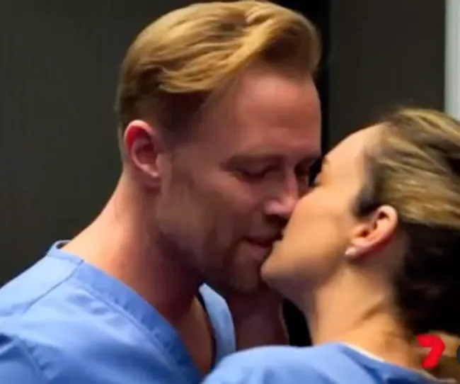 A man and woman in blue scrubs share a tender moment, leaning in for a kiss.