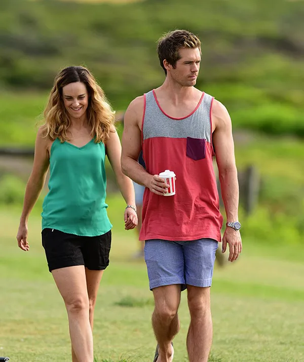 A man and woman walking on grass, the man holding a cup, both dressed casually under a sunny sky.