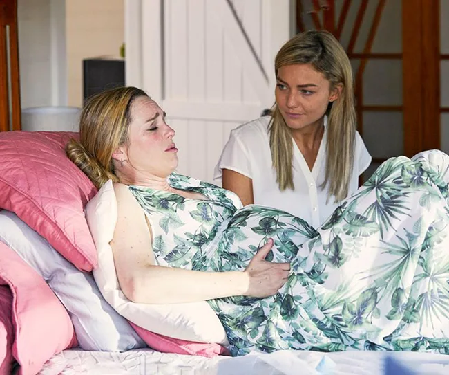 Pregnant woman in pain lying on bed with a concerned friend beside her in a floral-patterned dress.