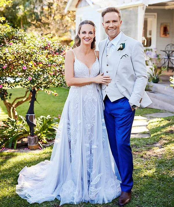 A couple in formal attire standing outdoors; woman in white gown, man in light suit with blue pants.