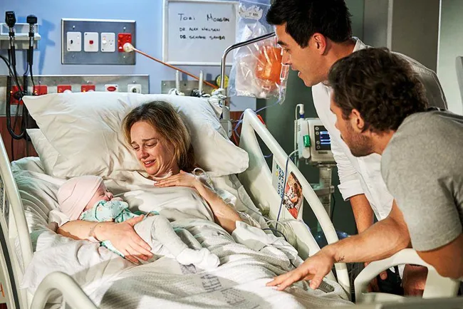 A woman in a hospital bed holding a baby, with two people standing beside her, in a medical setting.