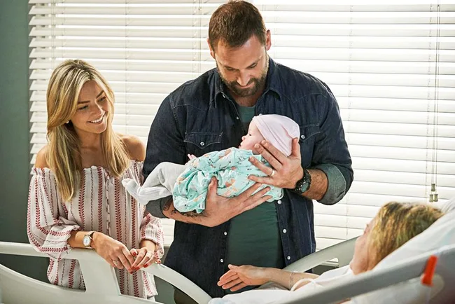 A man holds a newborn baby in a hospital room, with a smiling woman beside him and a person lying in bed.