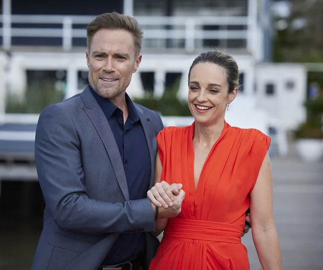 A man in a blue suit and a woman in a vibrant red dress smile while posing outside a modern building.