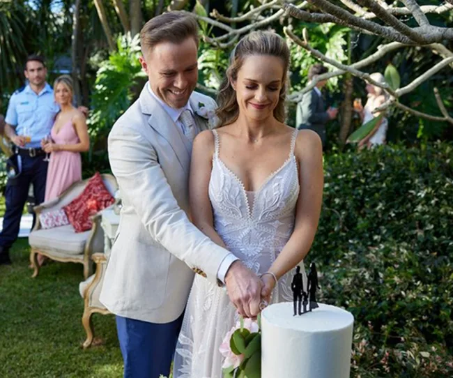 Bride and groom smiling as they cut a wedding cake in an outdoor garden setting.