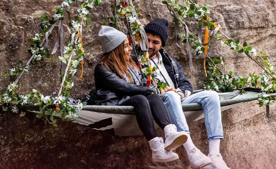 Couple sitting on a swing decorated with vines and flowers, smiling at each other.